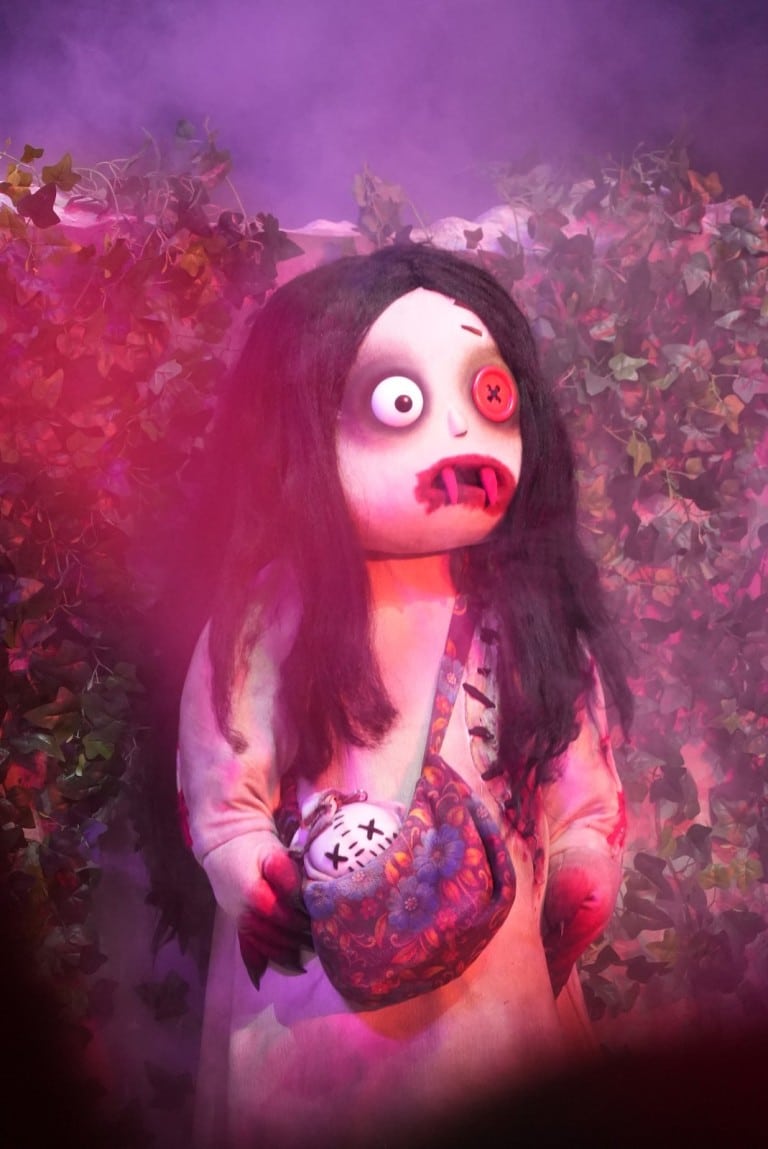 A costumed character with a large round head, long black hair, one red eye and fangs holds a round bag