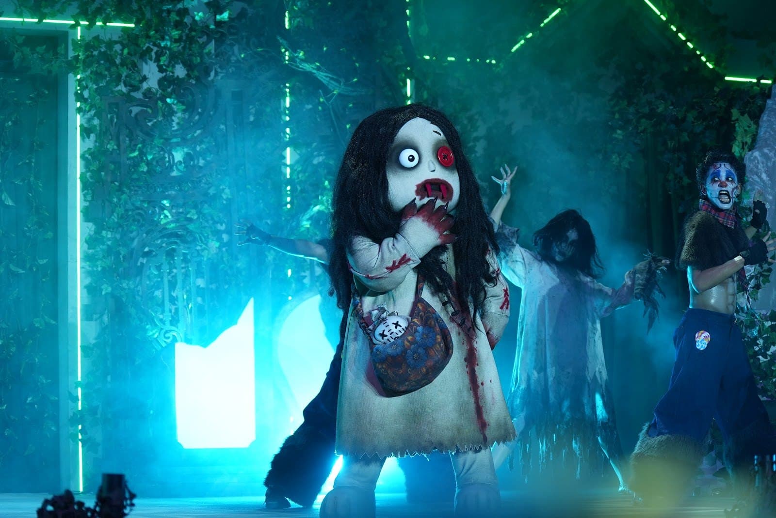 A costumed character with a large round head, long black hair and one red eye stands on a stage in teal light with dancers behind