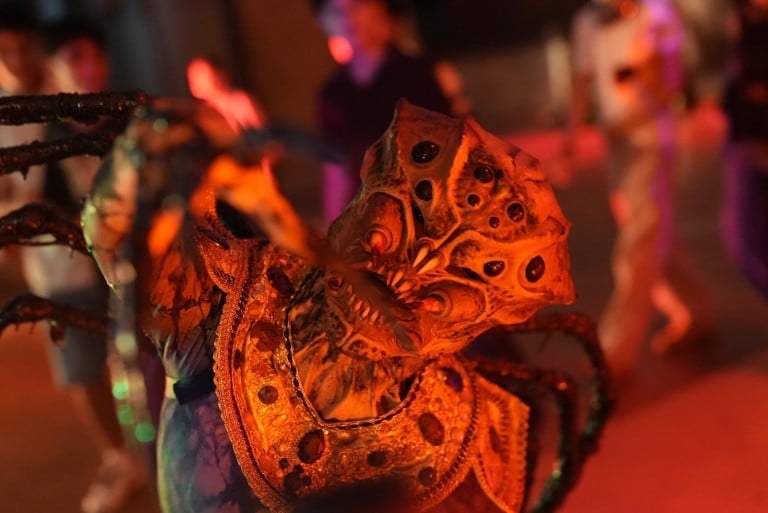 A performer in ornate layered armor with long spines crouches in orange light