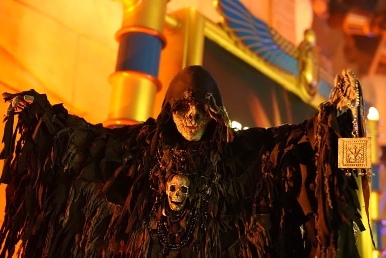 A performer in a tattered dark cloak and a skull mask spreads both arms in front of a temple wall
