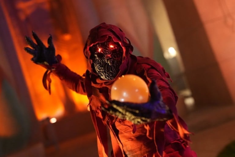 A performer in a red hood with glowing red eyes holds a glowing orb
