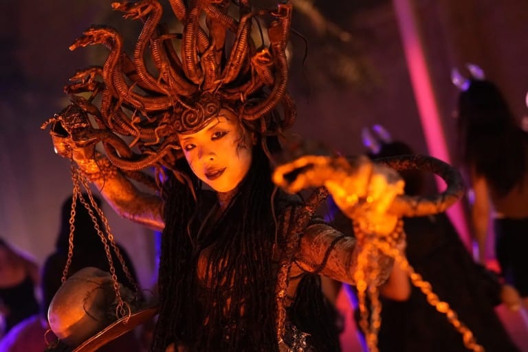 A performer with a headdress of coiled snakes and gold scale armor holds chains, lit orange