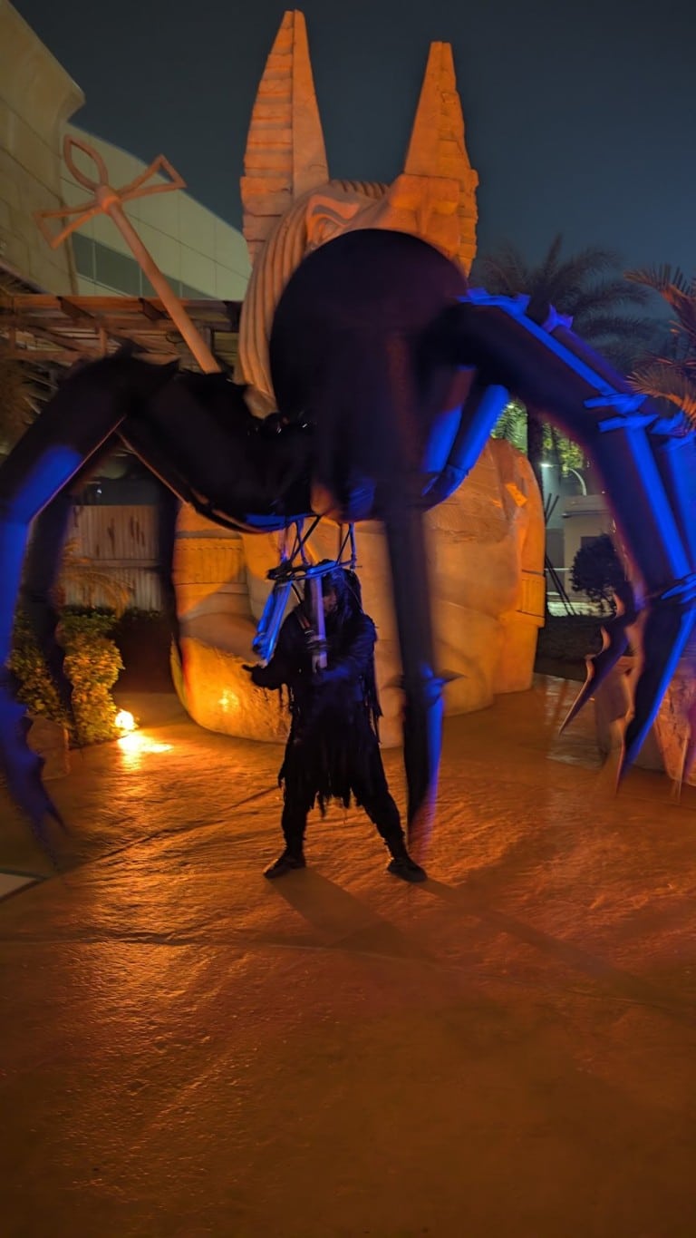 A performer walks under a giant black spider costume with long legs, lit blue, in front of a temple facade