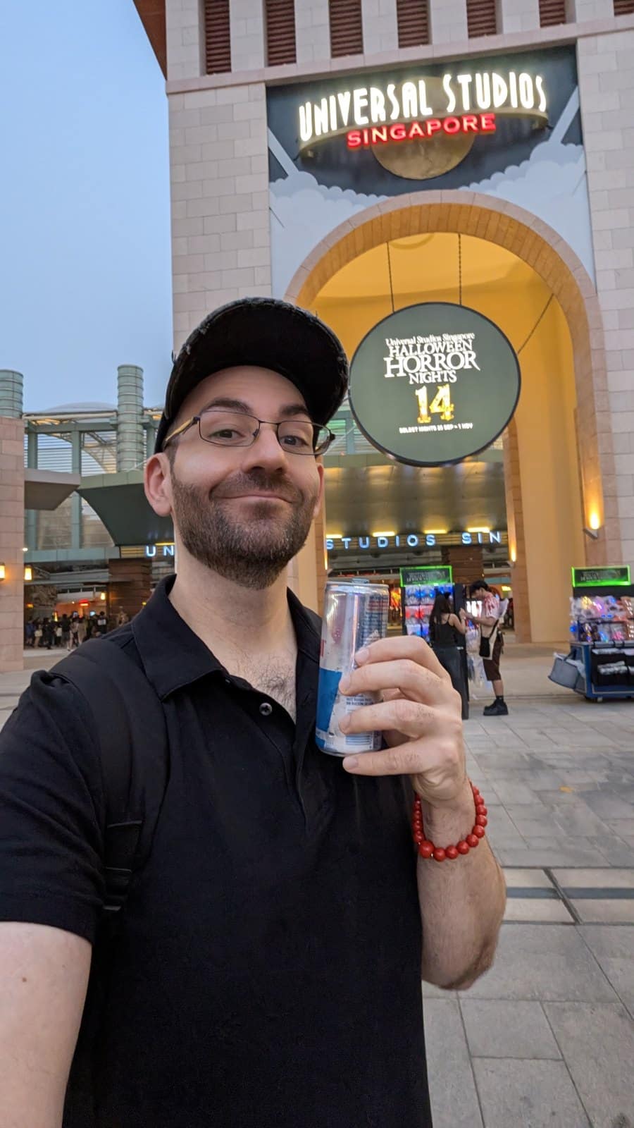 A man in a black cap and black shirt holds a drink can in front of the Universal Studios Singapore gate tower