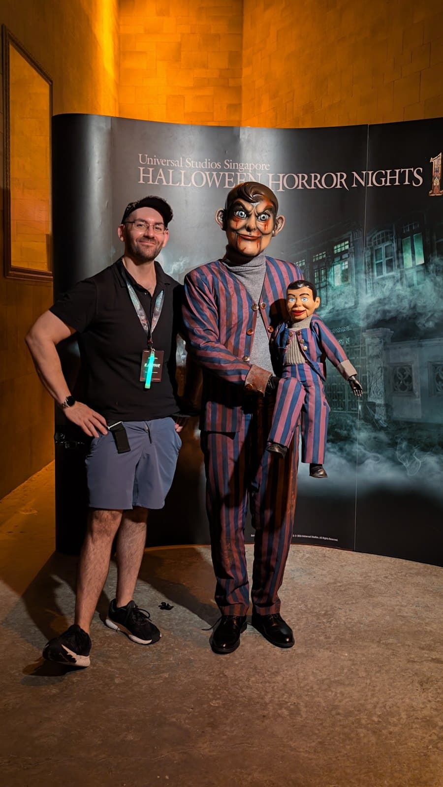 A man in a black shirt stands with arms crossed beside a costumed ventriloquist in a striped suit holding a matching dummy