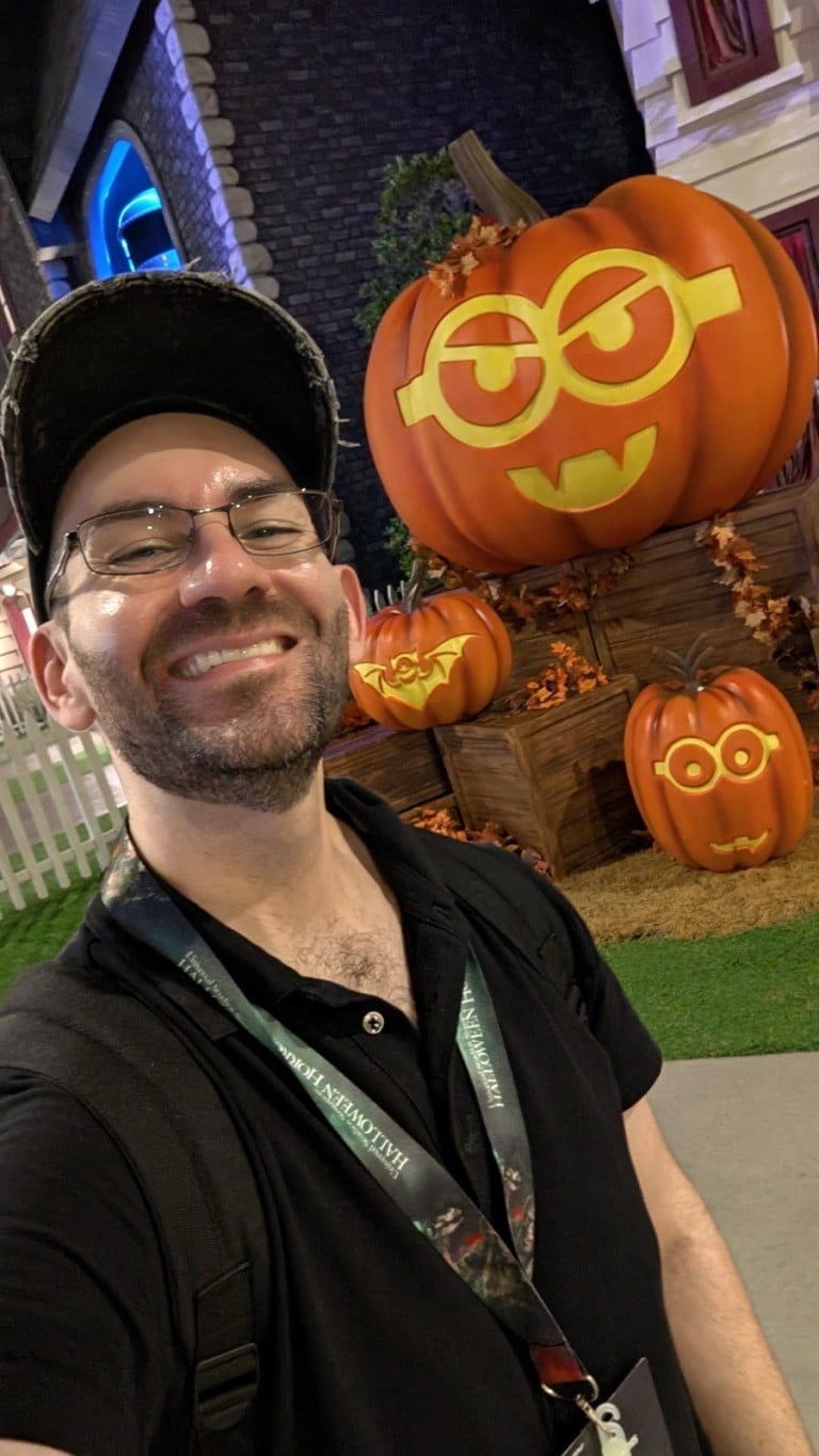 A man in a black cap smiles in front of a stack of jack-o'-lanterns carved with goggle-eyed faces