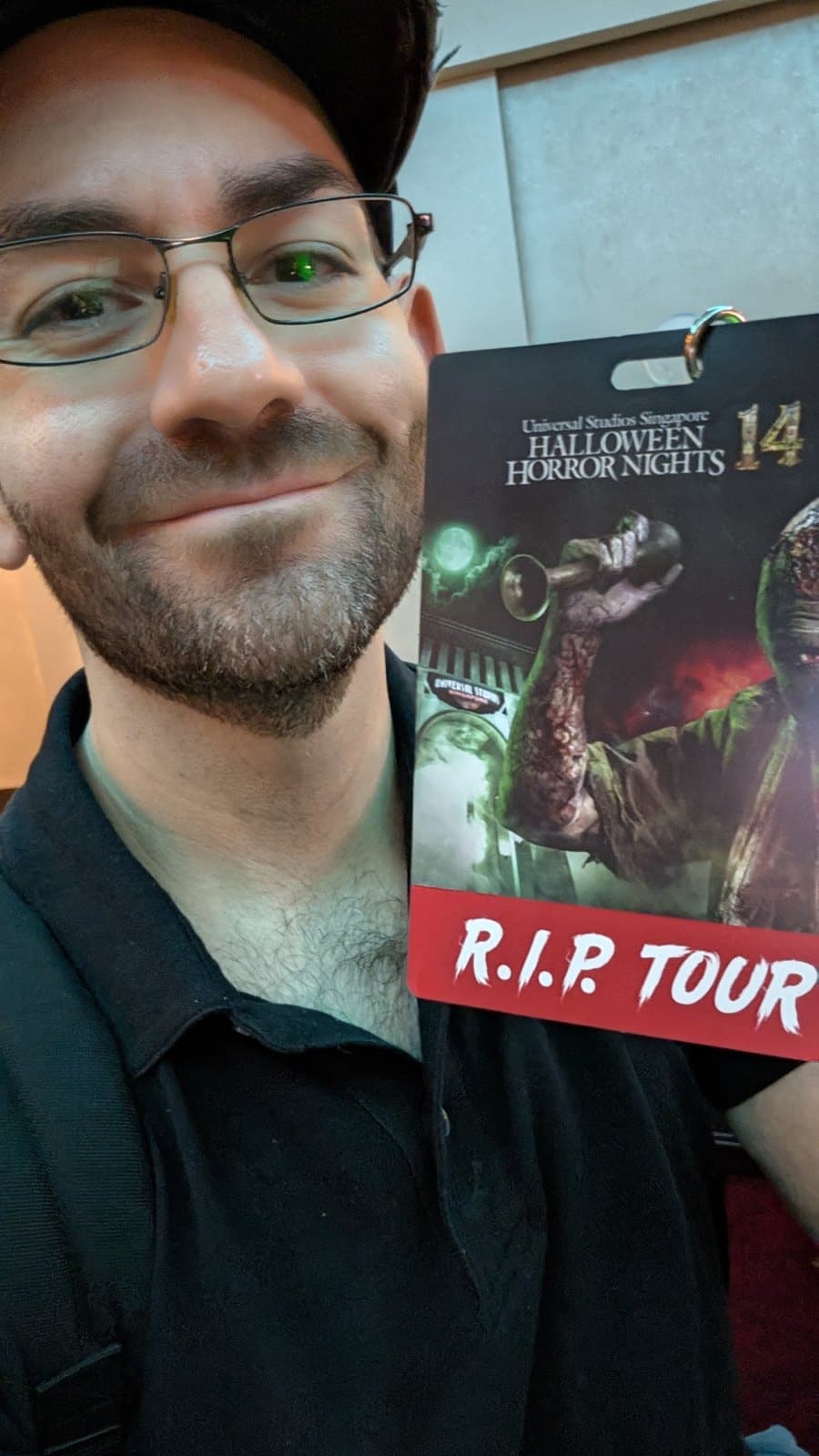 A man in glasses and a black cap holds up a lanyard card with a zombie illustration next to his face