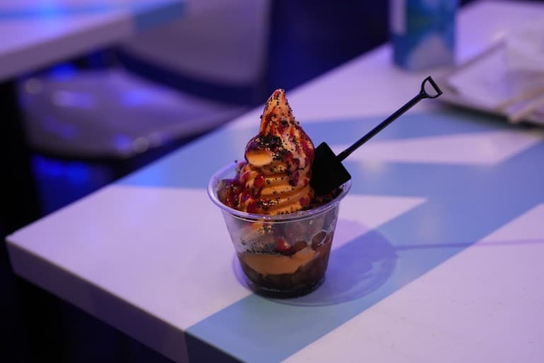 A cup of swirled soft serve with dark sauce and a small black shovel-shaped spoon on a white table