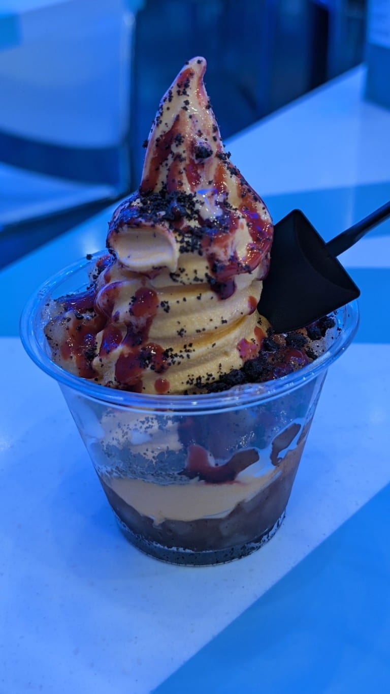 A cup of orange and white swirled soft serve with purple and dark toppings and a black shovel-shaped spoon