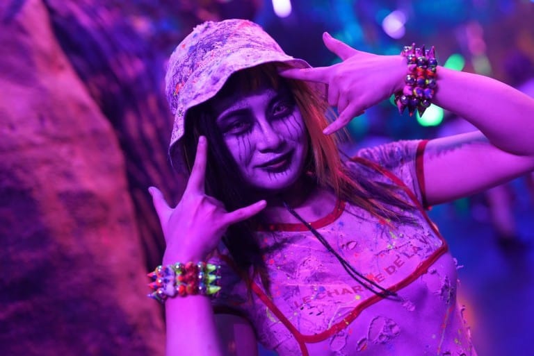 A performer in a bucket hat and pale face paint poses with both hands raised by her face in purple light