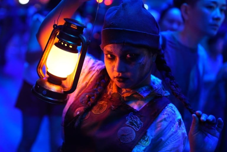 A performer in pale face paint, braids and a dark cap holds up a lit lantern in blue light
