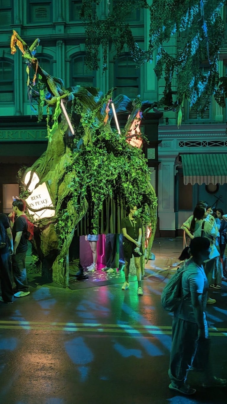 A twisted tree set piece wrapped in green and yellow light in the middle of a street, with people around it