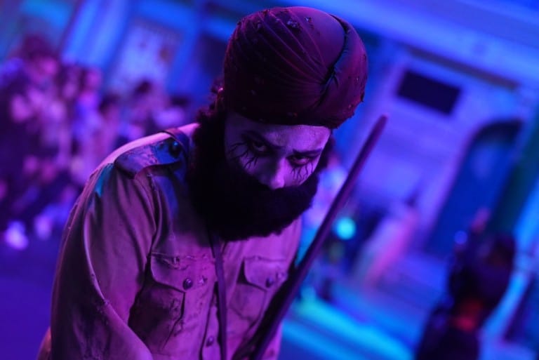 A performer in a purple turban and a long dark beard looks down in blue and purple light