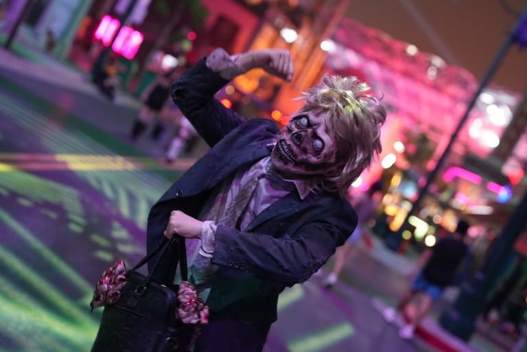 A performer in zombie makeup and a gray suit with wild gray hair poses with a handbag on a lit street