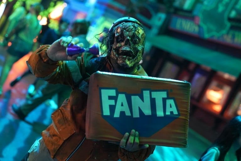 A performer in zombie makeup and a headscarf holds a board with a soft drink logo in teal light