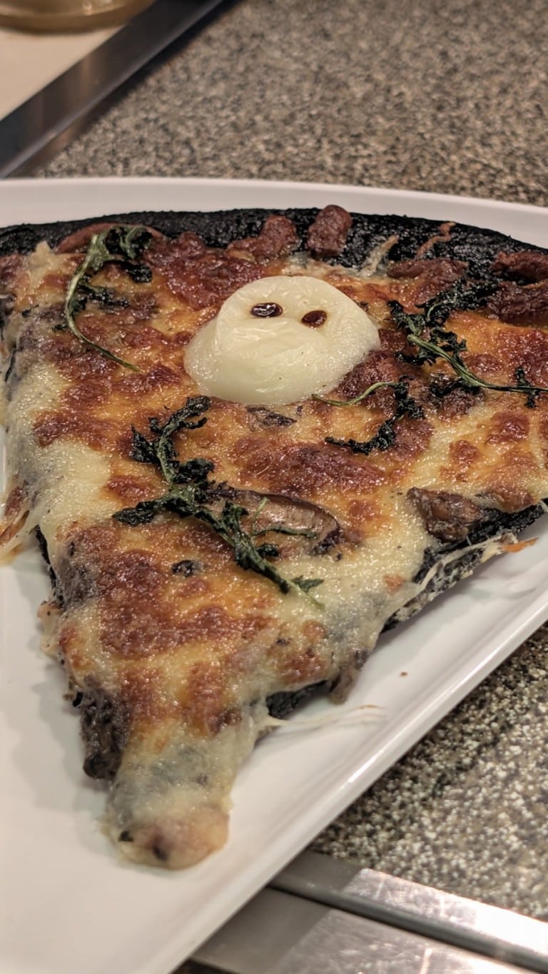 A slice of mushroom pizza with a white skull-shaped piece of cheese on top, on a white tray