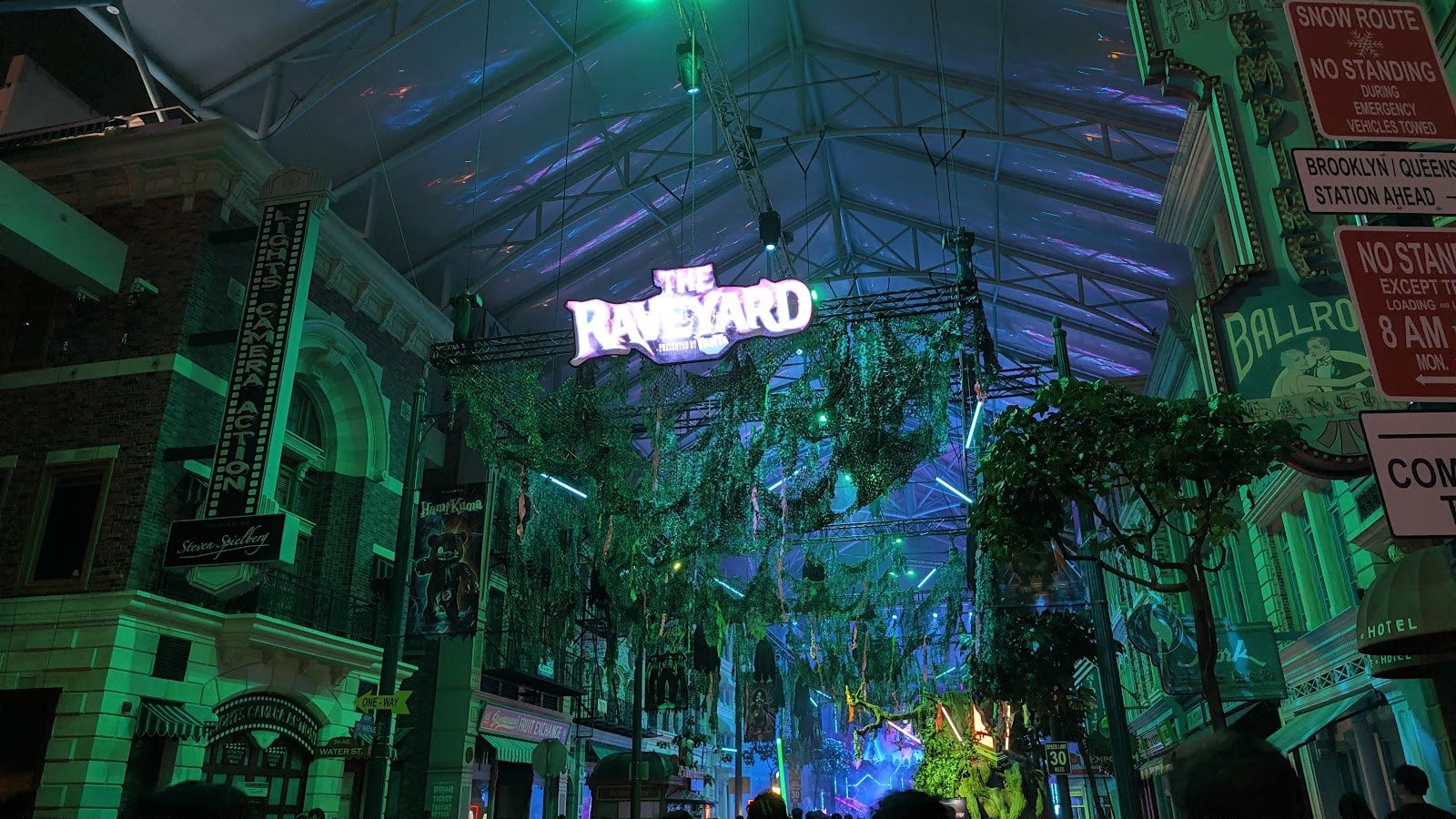 A glowing sign over a green-lit street of building fronts, with vines hanging from an overhead frame