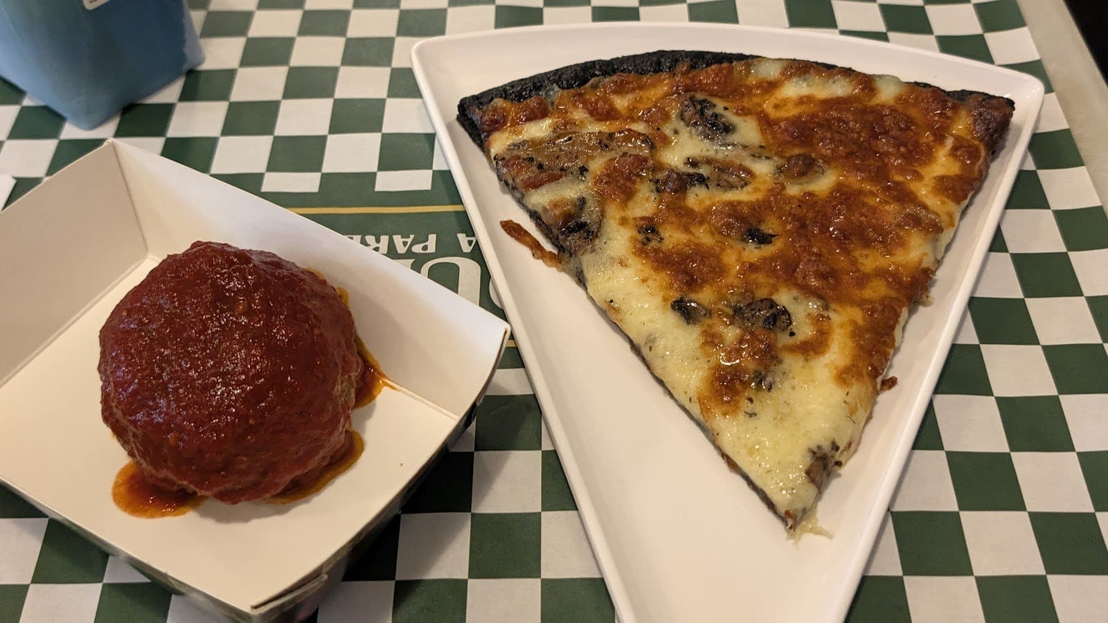 A slice of mushroom pizza on a white tray beside a meatball in red sauce in a paper boat, on a green checked table