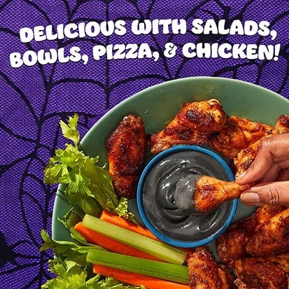 A chicken wing dipped into a bowl of black Hidden Valley Spooky Ranch, with celery and carrot sticks