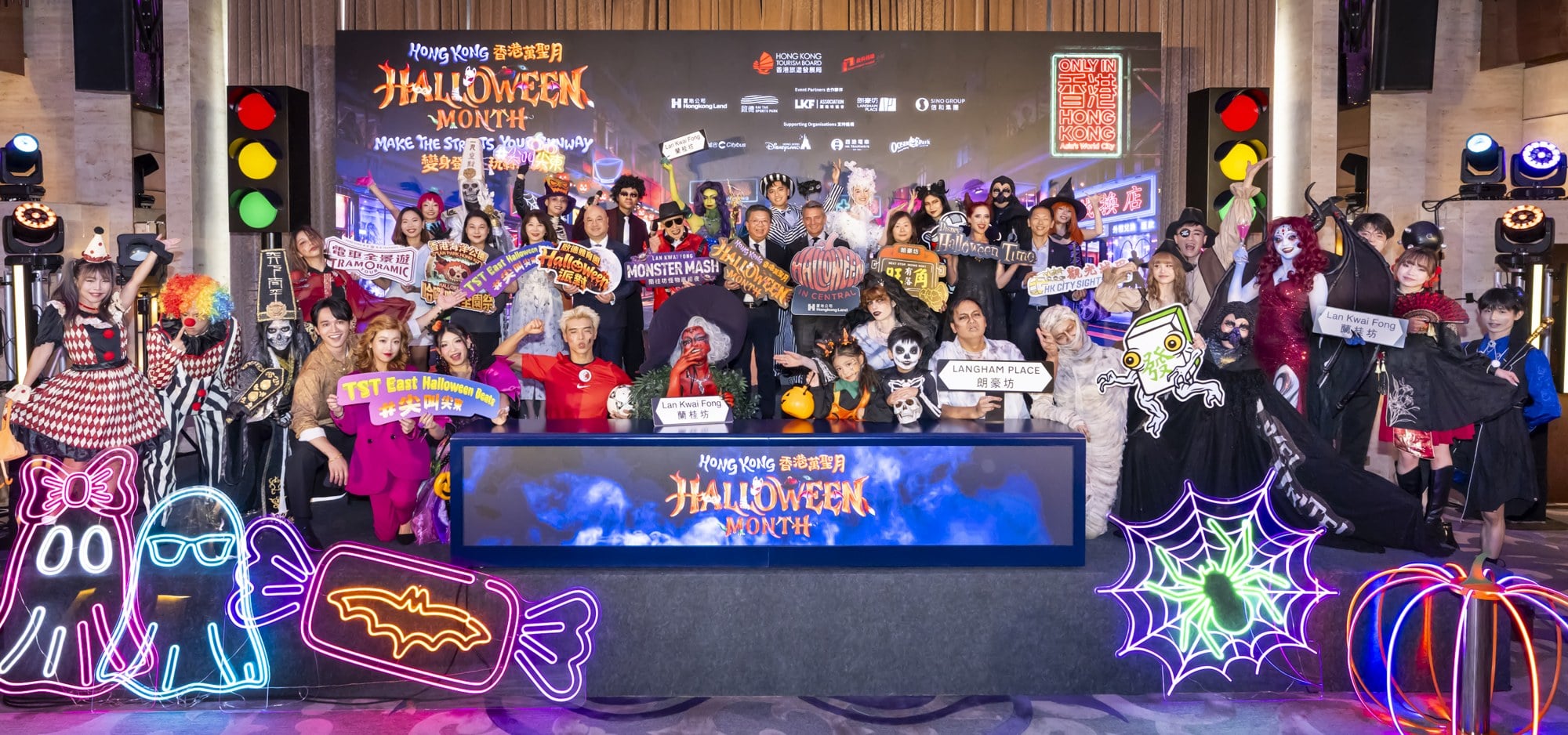Costumed performers and partner executives on stage at the Hong Kong Halloween Month announcement, holding signs for Lan Kwai Fong, Langham Place, and TST East Halloween Beats