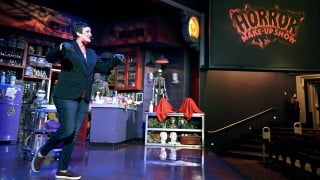 A performer in a dark blazer strides across a stage set with shelves of masks and prop heads, beside a screen showing a glowing Horror Make-Up Show sign