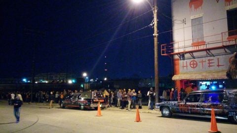 People line up at night outside a white building with red lettering that reads Hotel, with two painted hearses parked at the curb