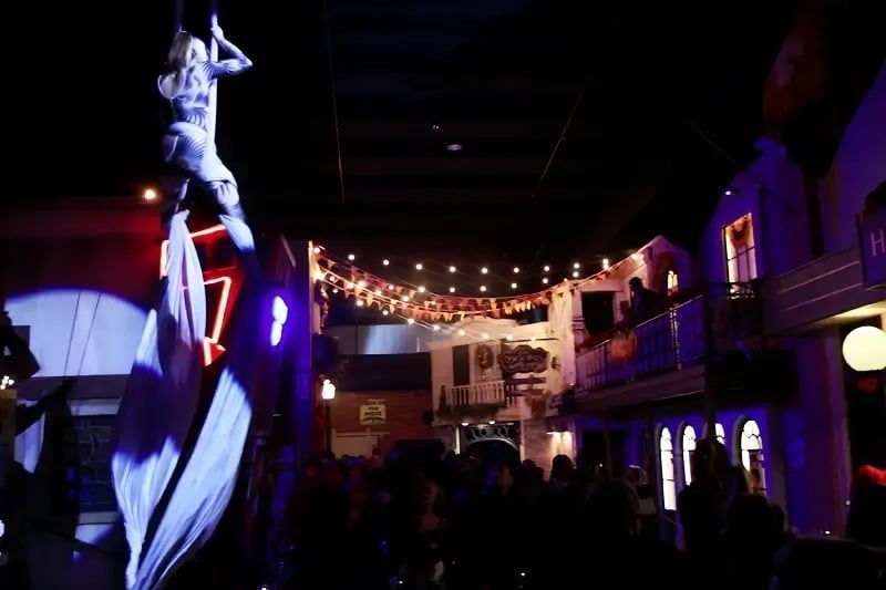An aerialist hangs from white silks above a crowd on an indoor street strung with lights