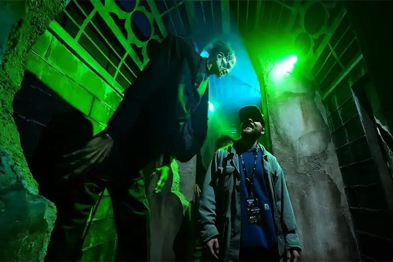 A tall scare actor leans over a guest in a green-lit stone corridor