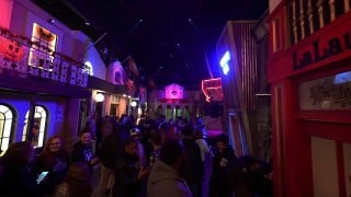 A crowd walks down an indoor street between two-story building facades lit in purple and red, with jack-o'-lantern decorations on a balcony