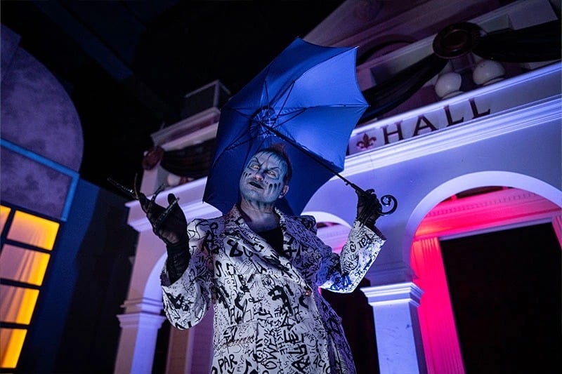 A scare actor in gray face paint and a black-and-white patterned suit holds a blue umbrella in front of a white building facade
