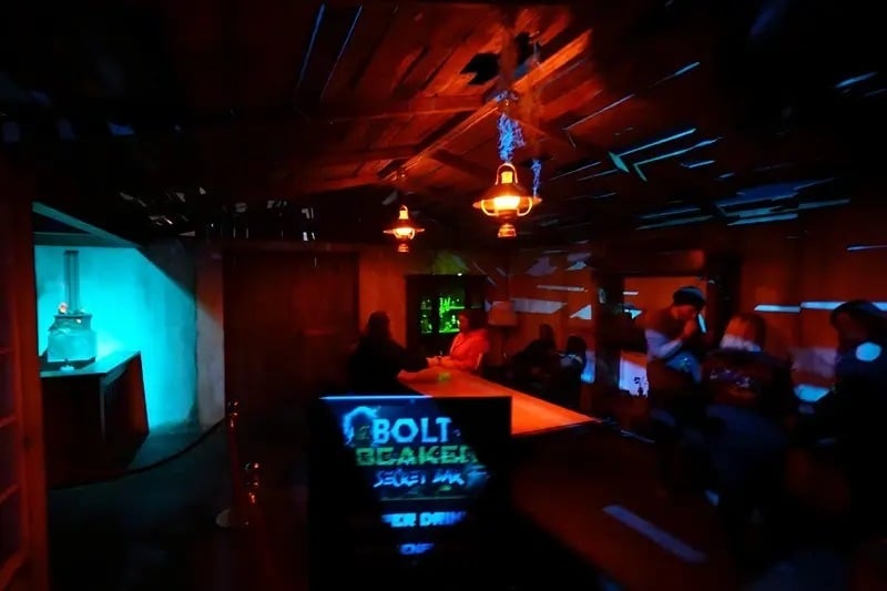 Guests sit along a bar in a dark room with a wood ceiling, red lantern light, and a glowing blue wall