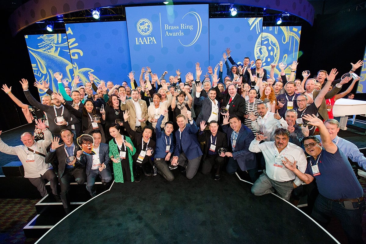A large group of award winners on a stage raising their hands and holding round glass trophies in front of a blue screen