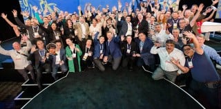 A large group of people on a stage raising their hands, several holding round glass trophies