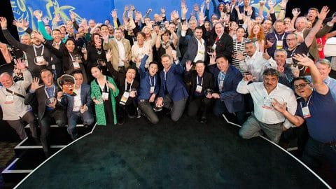 A large group of people on a stage raising their hands, several holding round glass trophies