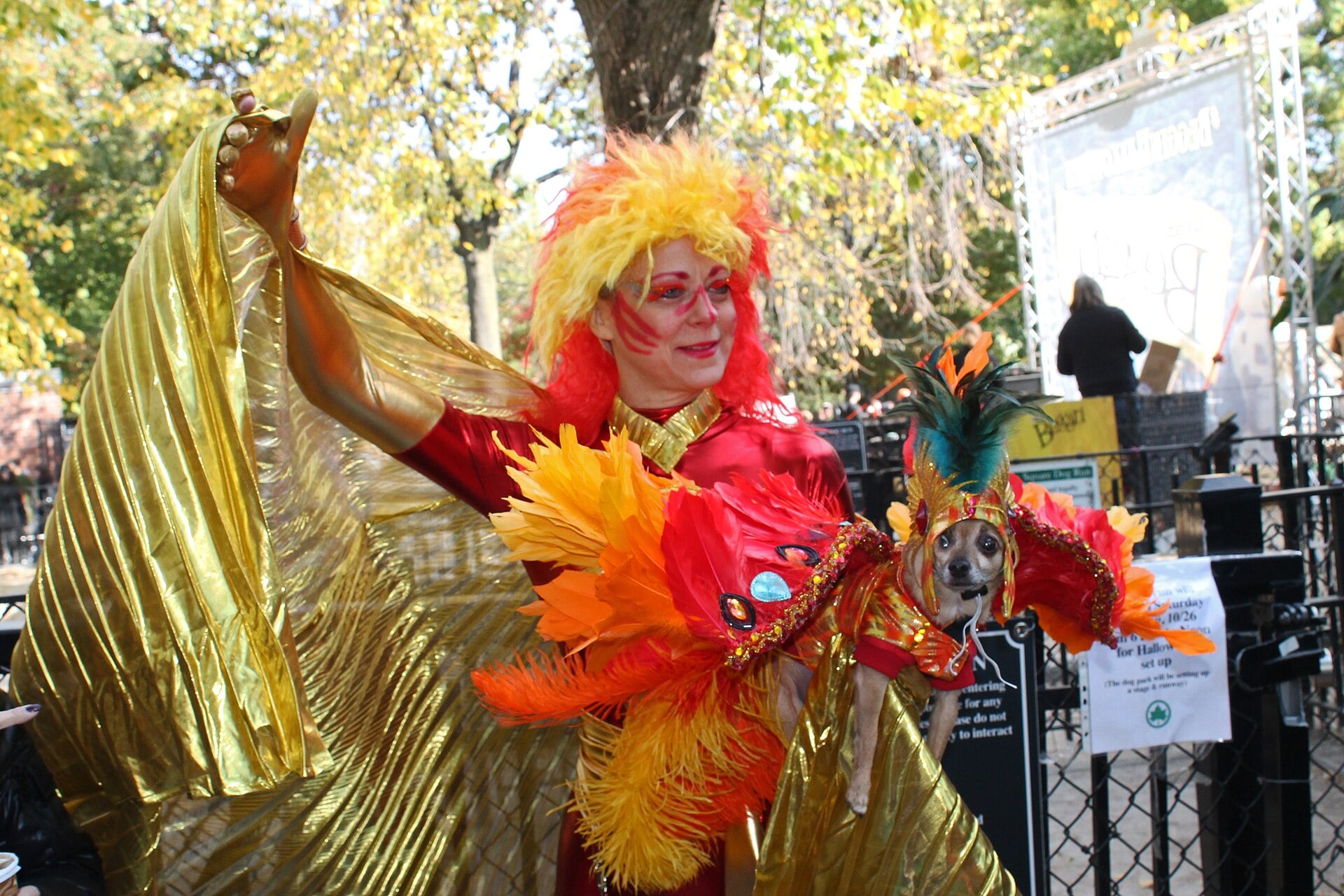 A dog in a feathered headpiece beside its owner in a red, orange, and gold firebird costume at the Tompkins Square Halloween Dog Parade.