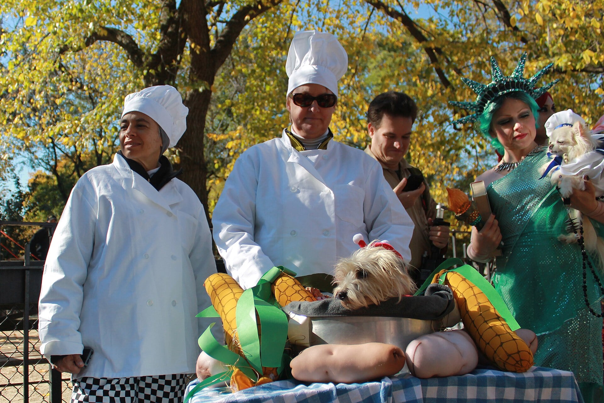 A small dog dressed as lobster stew sits in a pot, surrounded by people in chef whites and a Statue of Liberty costume.