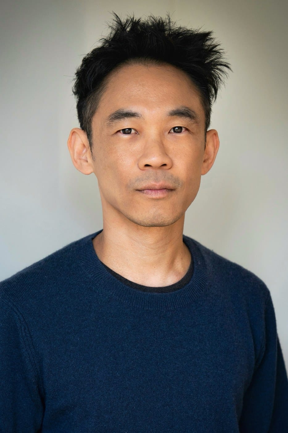 A man with short dark hair in a navy sweater looks at the camera against a pale gray wall