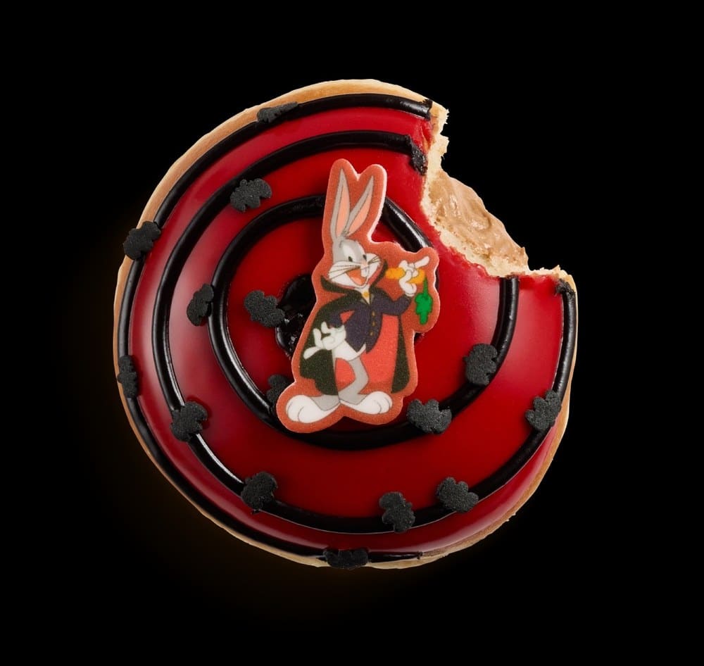 A doughnut with red icing, black drizzle, bat sprinkles, and a sugar piece of Bugs Bunny in a vampire cape, with a bite taken out