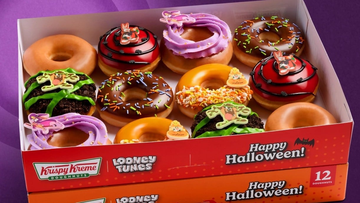 An open Krispy Kreme box of a dozen doughnuts topped with Looney Tunes character pieces, with Happy Halloween printed on the front of the box