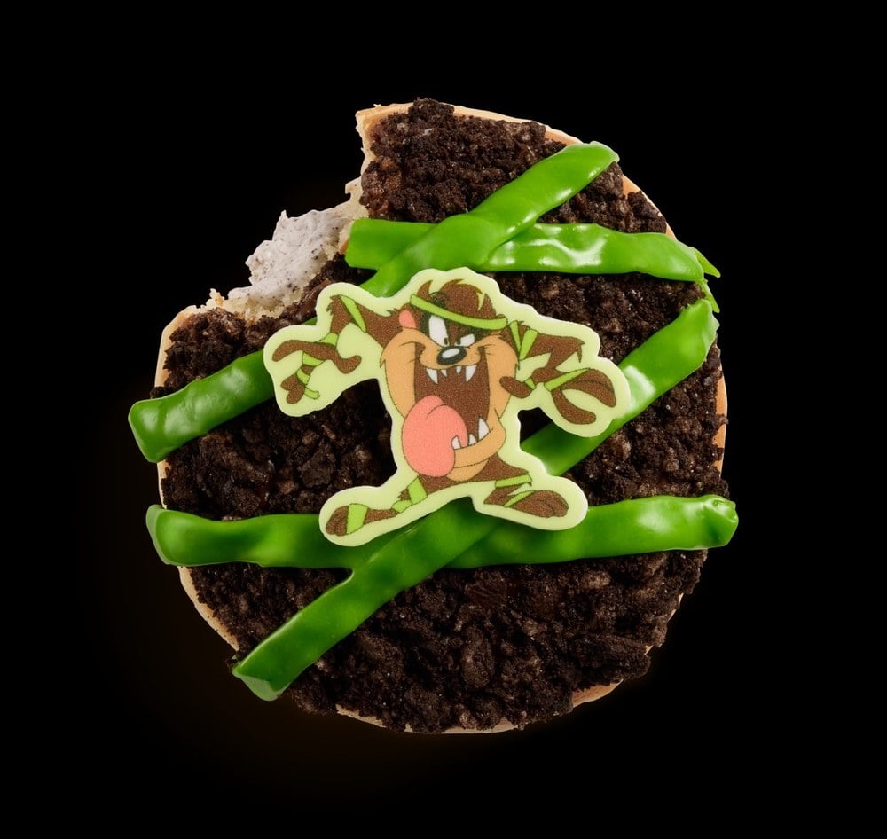 A doughnut covered in crushed cookie pieces with green icing stripes and a sugar piece of Taz wrapped as a mummy, with a bite taken out