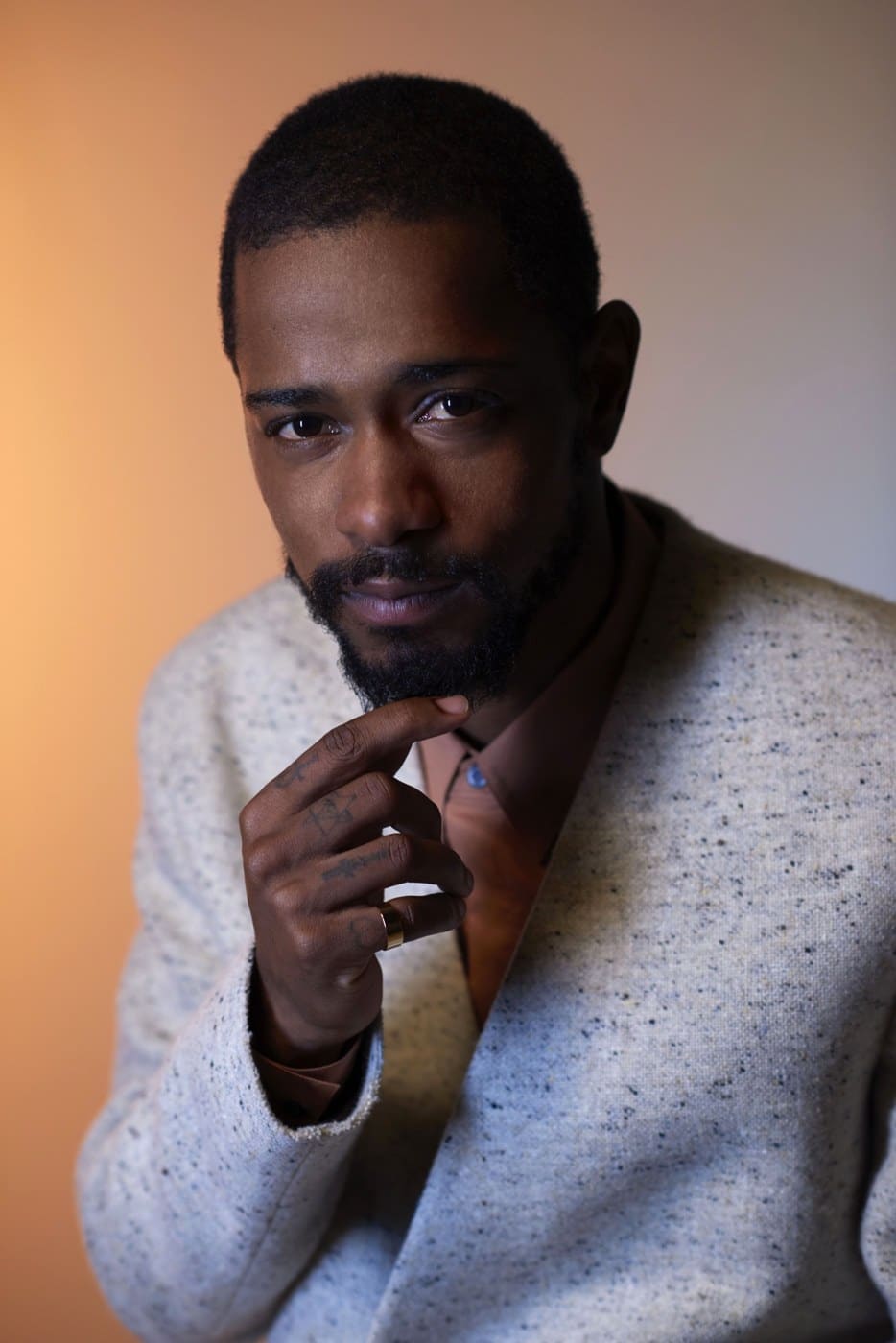 A man with a short beard in a cream speckled cardigan rests his chin on his hand against a warm tan background