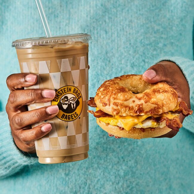 A person holds an Einstein Bros. iced coffee and a bacon, egg, and cheese bagel