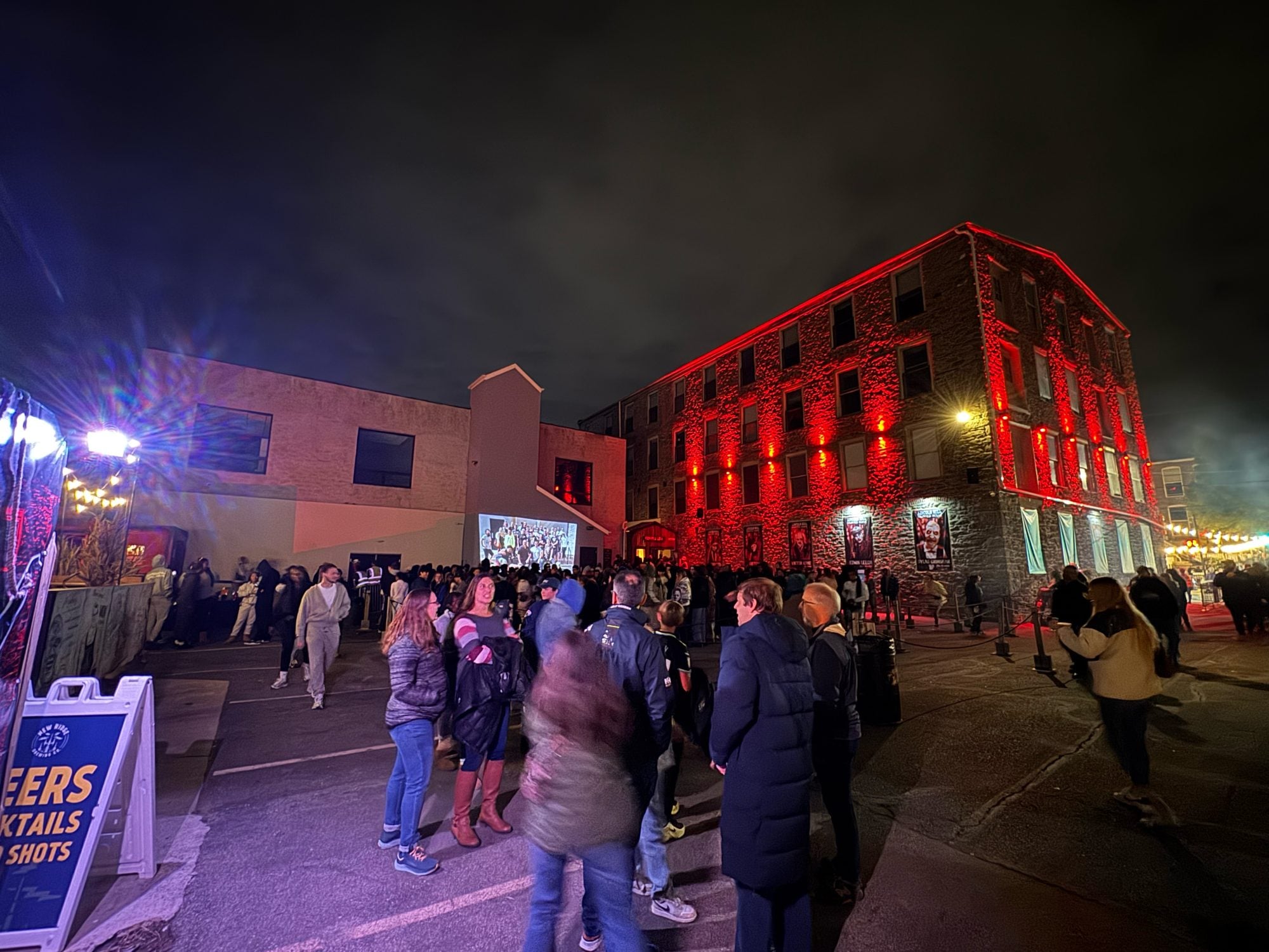 A crowd outside a red-lit stone mill building at night, with string lights and a lit screen on a wall