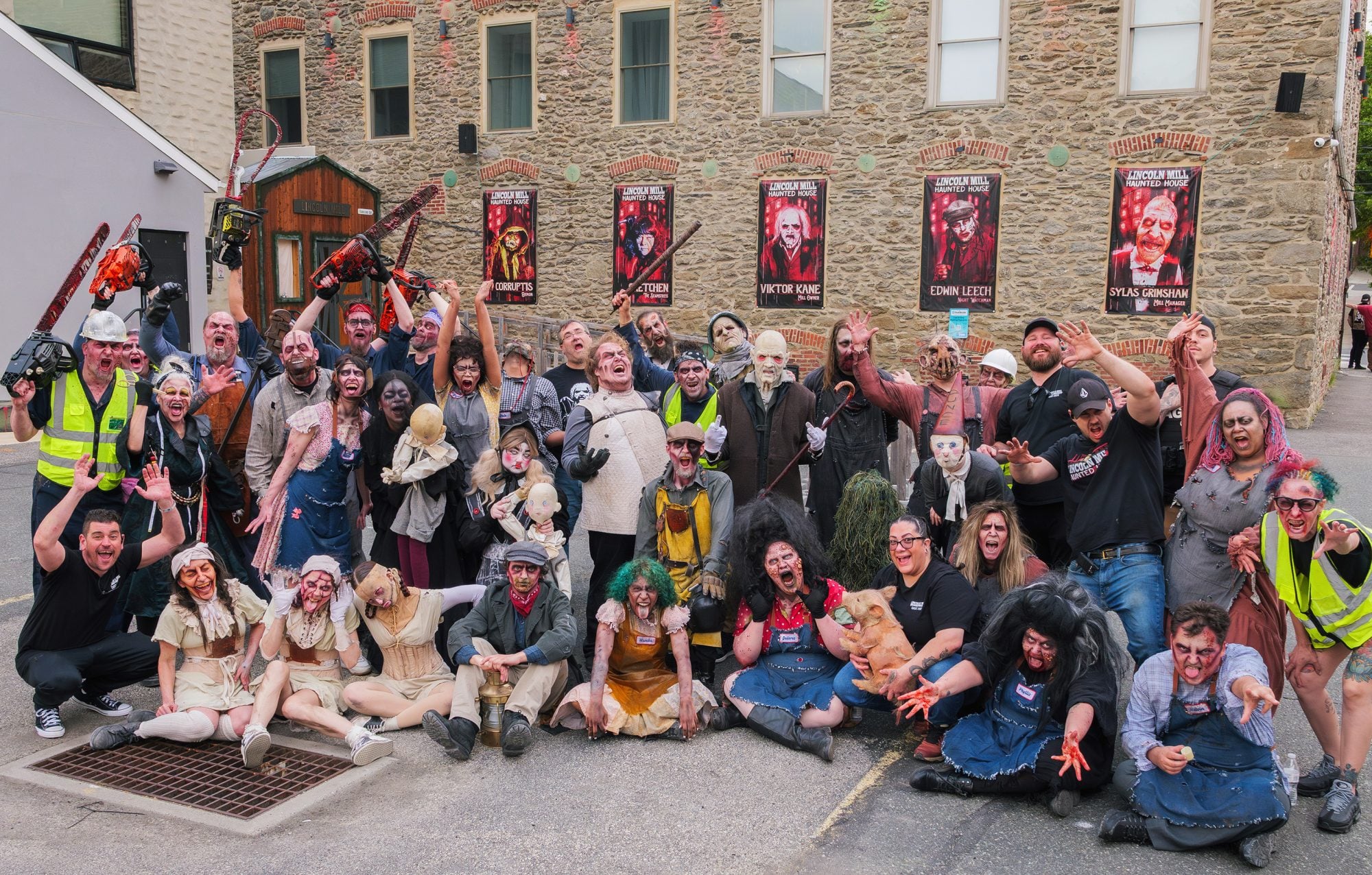 Dozens of costumed scare actors in horror makeup posing outside the stone mill, with chainsaws raised