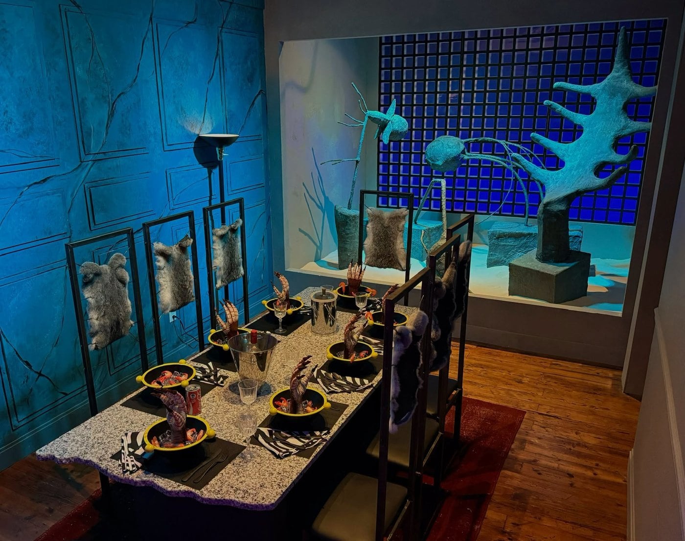 A dinner table with hands rising out of the bowls beside pale sculptures on a teal set, recreating the Beetlejuice dinner party