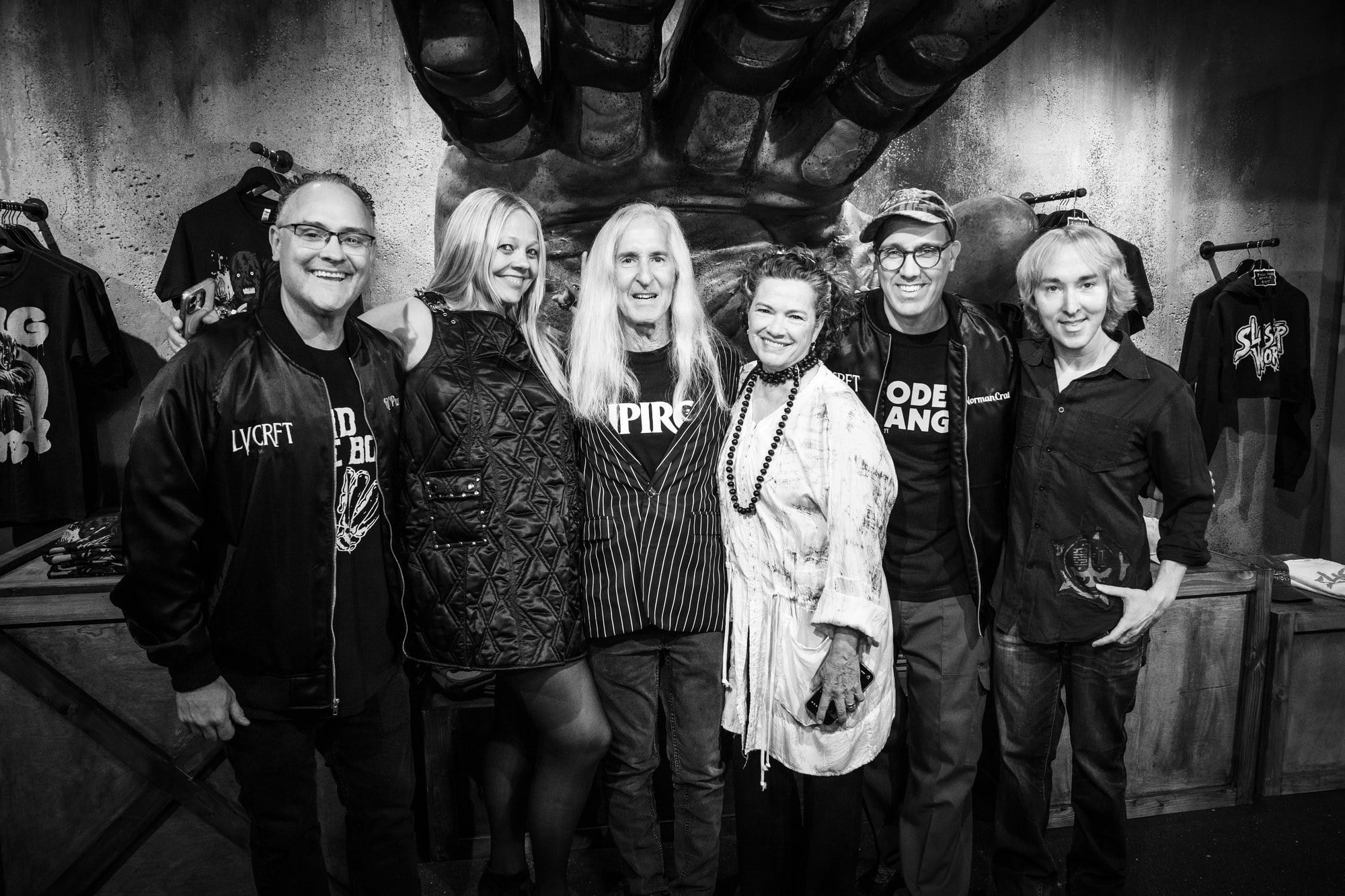 Six people stand together in a shop under a giant sculpted monster hand, in a black-and-white photo
