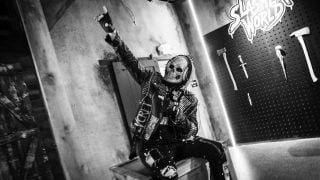 A person in a skull mask and a studded leather jacket raises a fist while sitting on a crate, in front of a wall of prop axes and knives, in a black-and-white photo