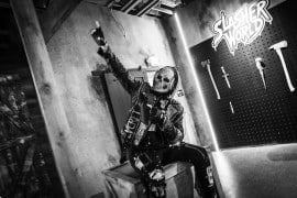 A person in a skull mask and a studded leather jacket sits on a crate with one fist raised, in front of a wall of prop axes and knives, in a black-and-white photo