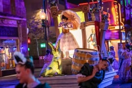 A performer in a pink dress holds a parasol on a float above an alligator, with dancers in maid and ballroom costumes around her