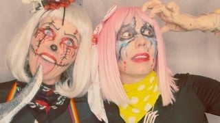 Mary, in a white wig with stitches painted across her face, licks a prop knife beside Margo, in a pink wig, as a fake severed hand rests on Margo's head